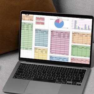 May include: A silver laptop displaying a colorful spreadsheet with charts and tables. The screen shows financial data, with various colored sections and a pie chart. The laptop is on a gray surface, with a brown pillow in the background.