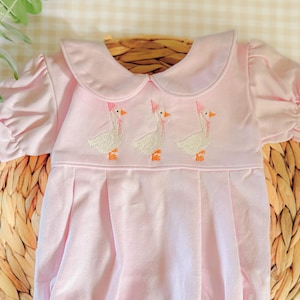 May include: A light pink baby romper with a Peter Pan collar and short, puffed sleeves. The yoke features an embroidered design of three geese wearing pink bonnets. The romper is gathered at the waist and has snaps at the bottom.