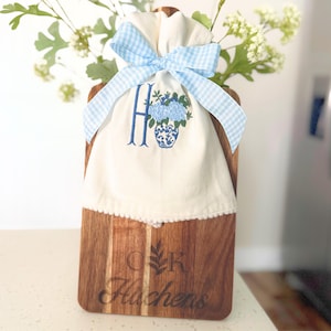 May include: A wooden cutting board with a white fabric bag featuring a blue embroidered floral design and the letter "H". The bag is tied with a light blue gingham bow. The board has engraved text and a floral design.
