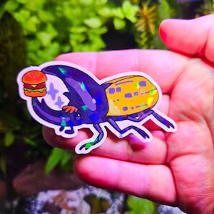 May include: A holographic sticker featuring a cartoon beetle holding a hamburger. The beetle has a purple head, yellow back, and purple legs. The hamburger is orange, yellow, and brown. The sticker has a shiny, iridescent finish.