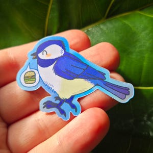 May include: A blue bird sticker with a white belly and a burger thought bubble. The bird has a smiling face and is outlined in a light blue color. The sticker is held in a person's hand.