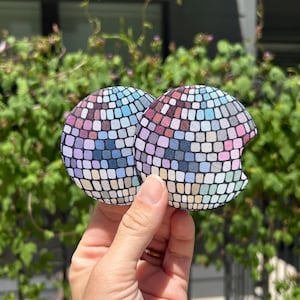 May include: Two circular car cup holder coasters featuring a multi-colored disco ball pattern with a grid of small squares in shades of blue, pink, purple, and white. One coaster has a small semi-circular notch cut out from the side edge.