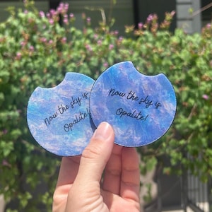 May include: Two circular car cup holder coasters featuring a blue and purple marbled pattern with a small semi-circular notch on the top edge. The text "Now the sky is Opalite!" is printed in black cursive lettering across the center of each coaster.