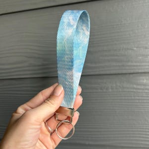 Puede incluir: Un llavero de muñeca teñido anudado azul y blanco con un cierre y un llavero de metal plateado. La tela tiene una apariencia texturizada. El llavero se sostiene en una mano sobre un fondo de madera gris.