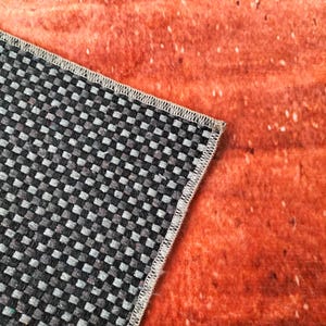 May include: Close-up of a section of a rug with a black and gray checkered pattern and a white stitched border. The rug is partially covering a textured orange surface with white speckles.