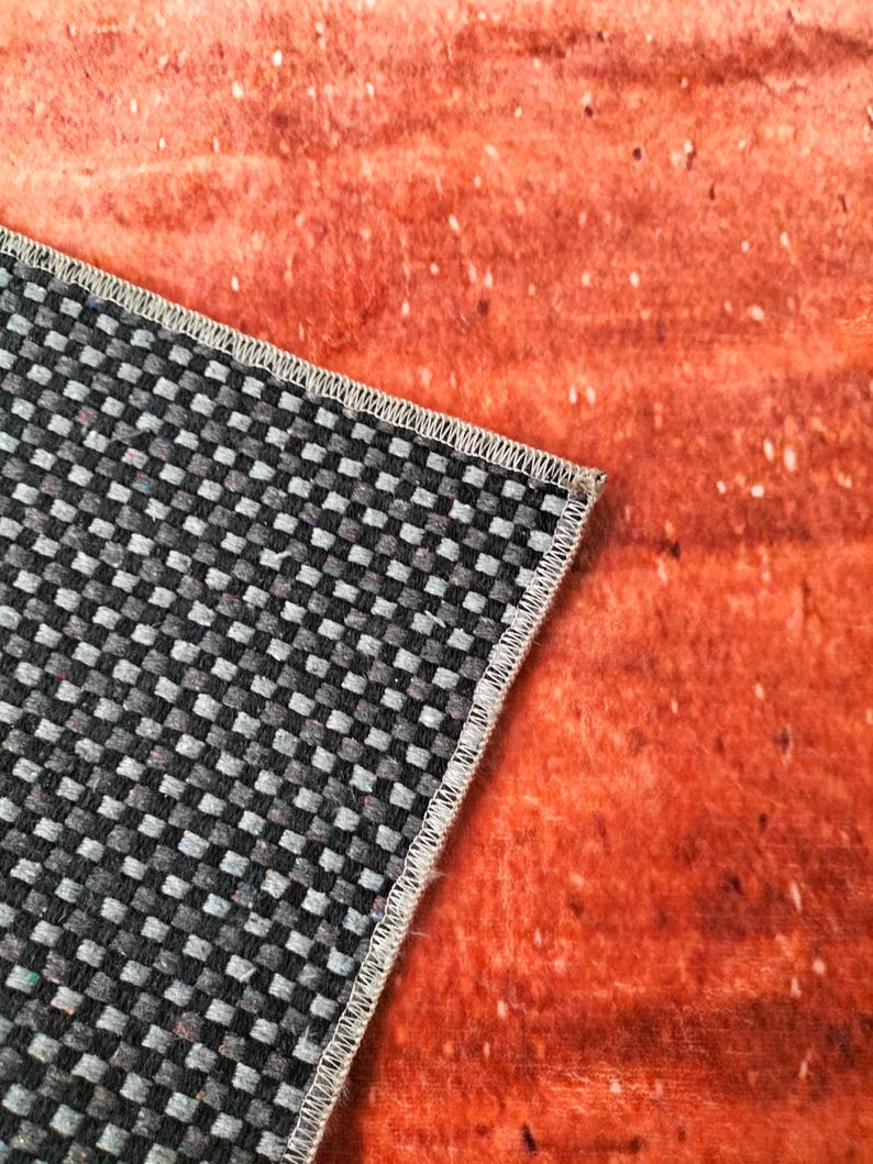May include: Close-up of a section of a rug with a black and gray checkered pattern and a white stitched border. The rug is partially covering a textured orange surface with white speckles.