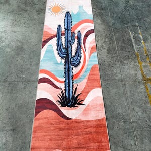 May include: A rectangular rug featuring a desert landscape design. A blue cactus is centered on the rug, with a sun and layered mountains in the background. The color palette includes shades of orange, red, blue, and white.