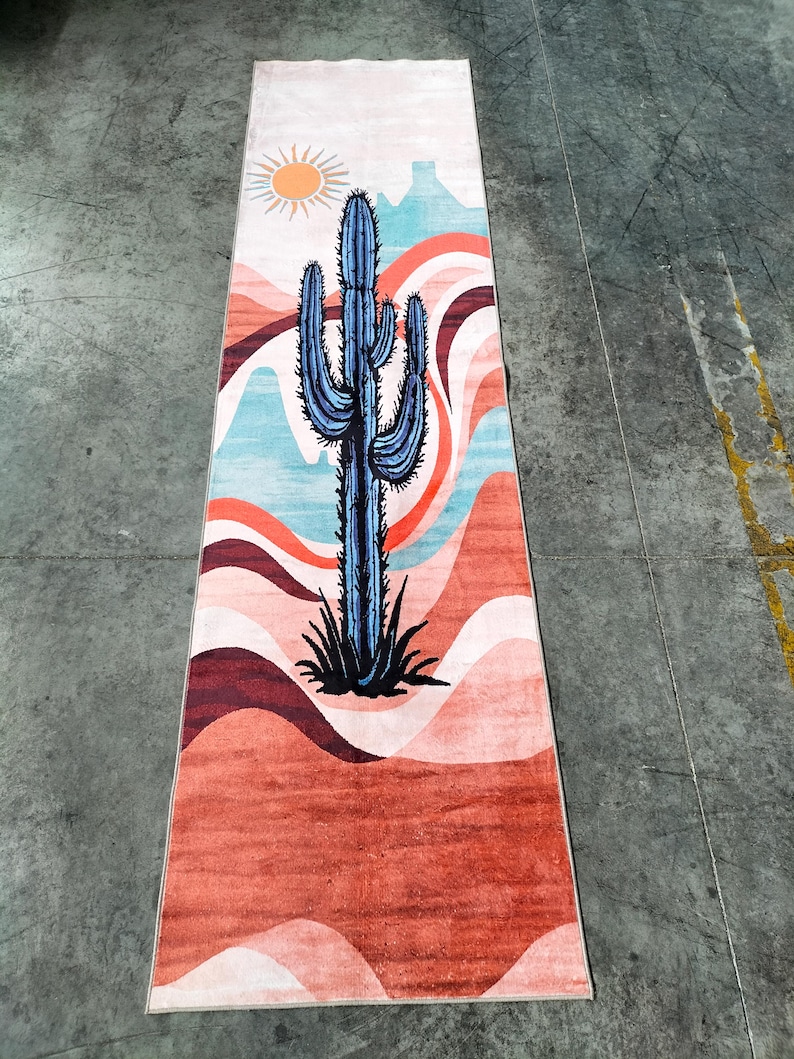May include: A rectangular rug featuring a desert landscape design. A blue cactus is centered on the rug, with a sun and layered mountains in the background. The color palette includes shades of orange, red, blue, and white.