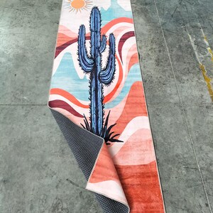May include: A rectangular rug with a desert-themed design. The rug features a large blue cactus, a yellow sun, and a landscape of red, orange, and blue wavy lines. The rug's underside is black with a white dot pattern.