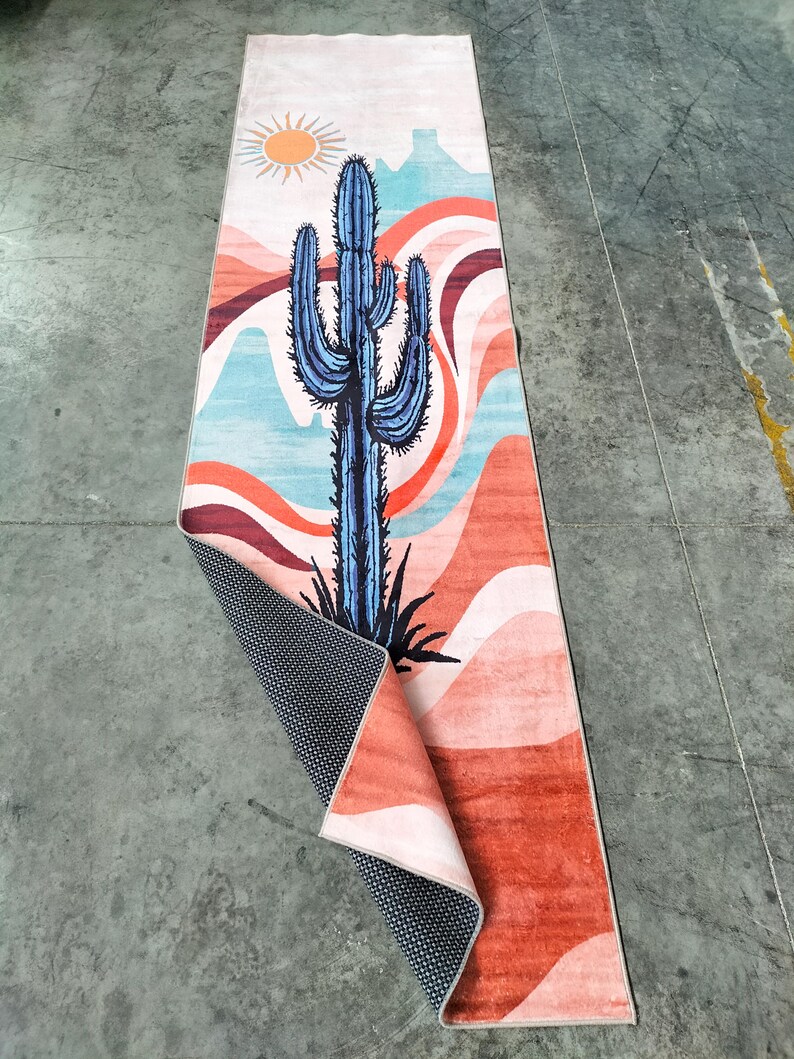 May include: A rectangular rug with a desert-themed design. The rug features a large blue cactus, a yellow sun, and a landscape of red, orange, and blue wavy lines. The rug's underside is black with a white dot pattern.