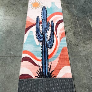 May include: A rectangular yoga mat featuring a desert landscape design. The mat showcases a blue cactus against a backdrop of orange, pink, and blue wavy patterns, with a sun and mountain silhouette. The mat's underside is a dark gray, textured material.