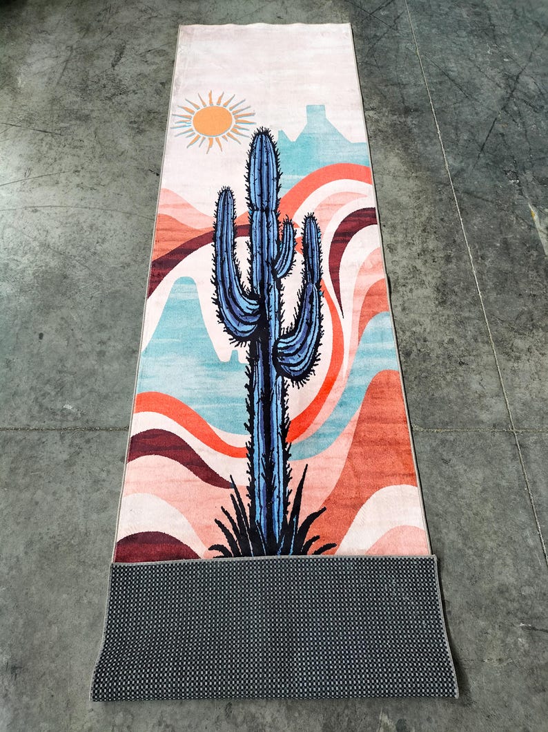 May include: A rectangular yoga mat featuring a desert landscape design. The mat showcases a blue cactus against a backdrop of orange, pink, and blue wavy patterns, with a sun and mountain silhouette. The mat's underside is a dark gray, textured material.