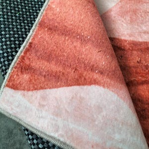 May include: Close-up of a folded rug with a watercolor-style design in shades of peach, coral, and white. The rug's edge is visible, showing a gray border and a black and gray checkered pattern underneath.