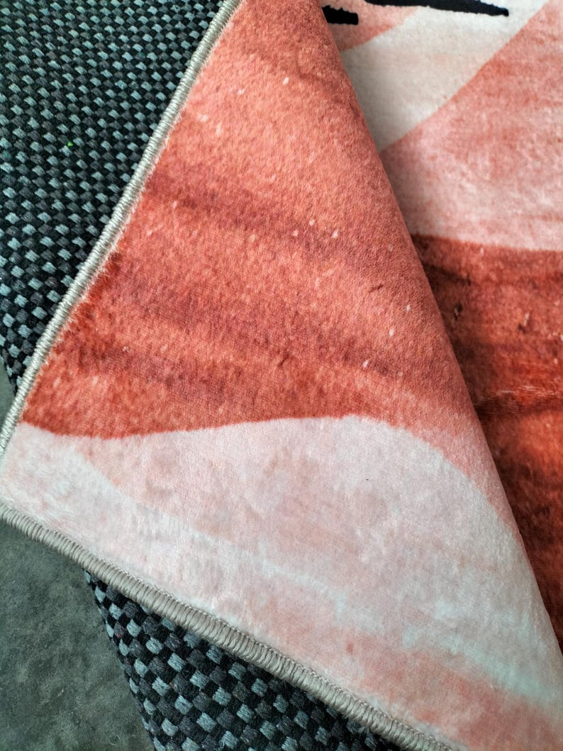 May include: Close-up of a folded rug with a watercolor-style design in shades of peach, coral, and white. The rug's edge is visible, showing a gray border and a black and gray checkered pattern underneath.