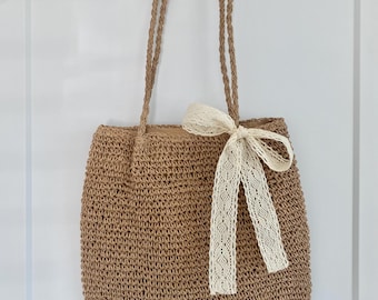 May include: A brown woven tote bag with a white lace ribbon tied in a bow. The bag has a woven strap.