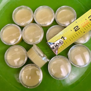May include: A close-up of ten small, clear containers filled with a light yellow substance. A yellow measuring tape is partially visible in the image.