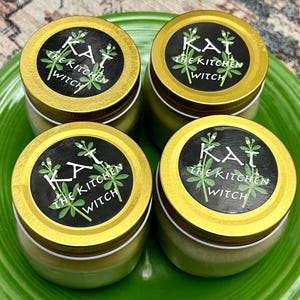 May include: Four glass jars with gold lids and white labels that read "KAT THE KITCHEN WITCH" in black text. The labels have a green plant design.