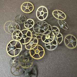 May include: A collection of various brass and metallic watch gears and wheels of different sizes and tooth counts, scattered in a cluster. These mechanical components feature central pinions and spokes, showcasing intricate metalwork and circular designs.