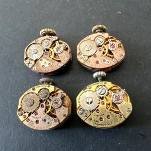 May include: Four vintage watch movements with exposed gears and jewels. The movements are in various shades of gold and copper, with intricate mechanical details. Some have text that reads "17 Jewels" and "Elgin 808 USA".