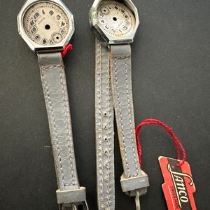 May include: Two silver-toned wristwatches with grey leather straps and a white face. The watches are missing the glass face and hands. A red tag with the text "Lamco Swiss Made" is in the bottom right corner of the image.