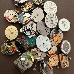 May include: A collection of assorted watch movements and watch faces in various shapes and sizes. The components feature metallic, white, and green surfaces with visible gears, quartz mechanisms, and printed text including Geneve, Fossil, and Gianello.