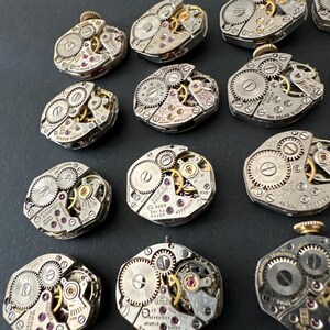 May include: A collection of vintage watch movements, showcasing intricate gears, jewels, and metal components. The silver-toned mechanisms feature visible cogs, screws, and ruby-colored jewels. Some movements have text that reads "GRUEN WATCH CO" and "17 JEWELS".