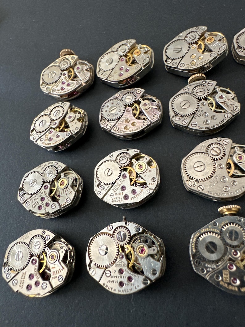 May include: A collection of vintage watch movements, showcasing intricate gears, jewels, and metal components. The silver-toned mechanisms feature visible cogs, screws, and ruby-colored jewels. Some movements have text that reads "GRUEN WATCH CO" and "17 JEWELS".