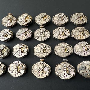 May include: A collection of twenty vintage watch movements, each with intricate gears, jewels, and metal components. The watch mechanisms are silver-toned with gold and red accents, arranged on a dark surface.