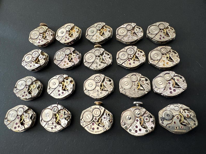 May include: A collection of twenty vintage watch movements, each with intricate gears, jewels, and metal components. The watch mechanisms are silver-toned with gold and red accents, arranged on a dark surface.