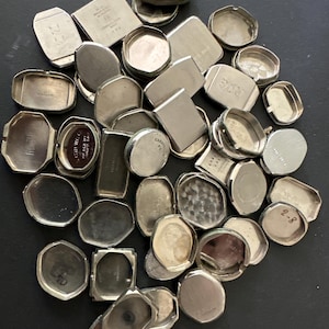 May include: A collection of vintage watch cases in various shapes and sizes, including round, rectangular, and square. The cases are silver-toned and appear to be made of metal. Some cases have visible markings and engravings.