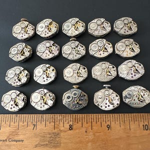 May include: A collection of twenty-one vintage watch movements, each with intricate gears, jewels, and metal components. The movements are silver and gold in color, arranged on a black surface. A wooden ruler with inch markings is placed below the watch movements.