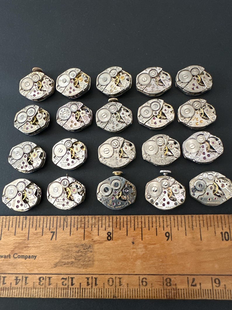 May include: A collection of twenty-one vintage watch movements, each with intricate gears, jewels, and metal components. The movements are silver and gold in color, arranged on a black surface. A wooden ruler with inch markings is placed below the watch movements.