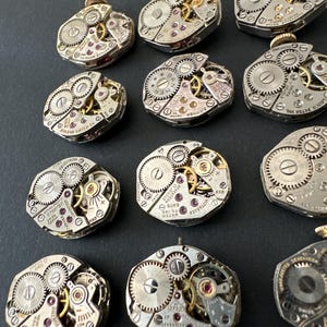 May include: A collection of vintage watch movements, showcasing intricate gears, jewels, and metallic components. The open-faced mechanisms display a variety of shapes and sizes, with visible text and details. The watch parts are arranged on a dark surface.