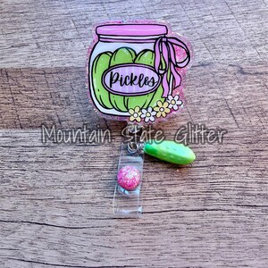 May include: A pink and green pickle jar badge reel with the word "Pickles" on the jar. The badge reel has a pink glitter ball and a green pickle charm. The text "Mountain State Glitter" is at the bottom.