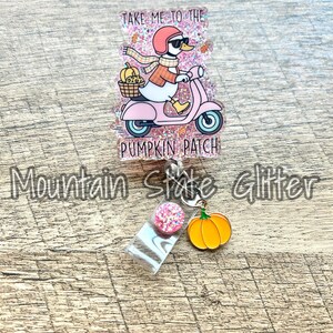 May include: A glittery badge reel with a duck riding a pink scooter, carrying pumpkins in a basket, and wearing a helmet and scarf. Text reads "Take Me To The Pumpkin Patch". Includes a pink glitter ball and a pumpkin charm.