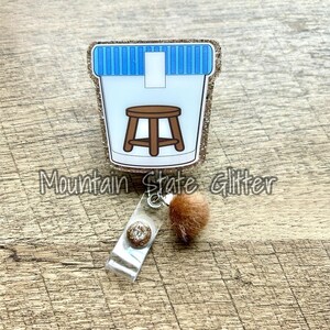 May include: A gold glitter badge reel with a clear plastic charm featuring a small brown stool inside a blue and white striped container. The charm is attached to a brown fuzzy pom-pom.