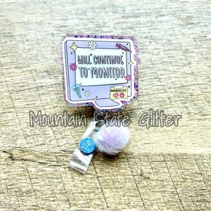 May include: A purple and pink glitter badge holder with a white screen that says "Will Continue To Monitor" and a yellow speech bubble that says "#NurseLife". The badge holder has a blue and white glitter pom-pom attached to a silver clip.