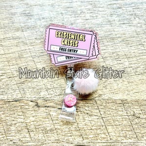 May include: A pink badge reel with the text "Existential Crisis" and "Free Entry" on a ticket-shaped design. It features a fluffy pink pom-pom and a small, glittery pink circle. The background is a light brown wood grain.