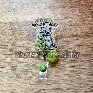 May include: A badge reel with a raccoon holding a jar of pickles. The text "Pickles and Panic Attacks" is above the raccoon. The reel has a green pom-pom and a green glitter button.