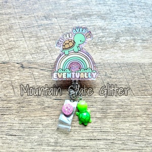 May include: A retractable badge reel with a turtle design. The reel features a turtle on a rainbow with the text "I'll get over it eventually". A small pink glitter circle and a green turtle charm are attached.