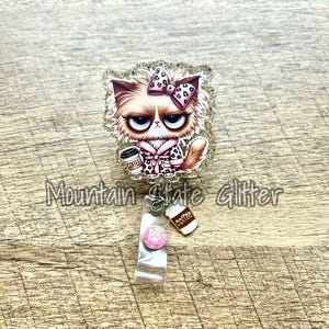May include: A glitter-covered cat-shaped badge holder with a pink bow, holding a coffee cup that says "Coffee". The badge holder is attached to a clear plastic retractable badge reel with a pink glitter charm.
