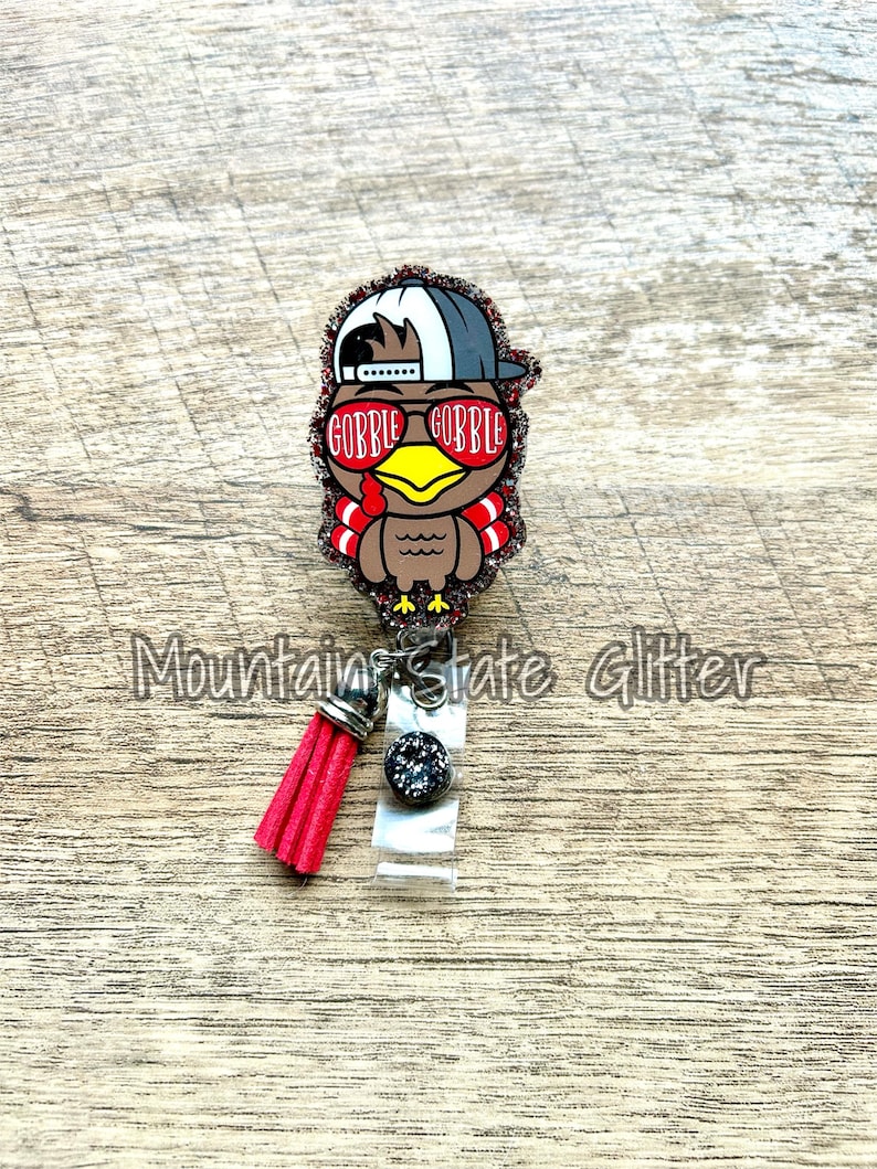 May include: A glitter-covered turkey wearing a backwards baseball cap and sunglasses with the text "Gobble Gobble" on the sunglasses. The turkey is attached to a red tassel and a clear badge holder.