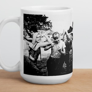 Peut inclure: Mug en céramique blanche avec une image en noir et blanc d'un groupe de personnes dansant en extérieur. La tasse a une grande anse et est posée sur une surface en bois.