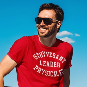 Puede incluir: Un hombre con una camiseta roja con texto blanco que dice "Stuyvesant Leader Physical Ed" y pantalones vaqueros azules. Sonríe y lleva gafas de sol. Está de pie frente a una barandilla de metal.