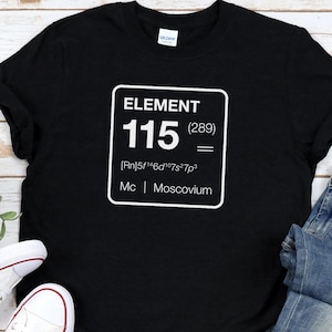 May include: Black t-shirt with a white graphic of Element 115, Moscovium, and its atomic number. The shirt is paired with blue denim jeans and white canvas sneakers with red and blue accents.
