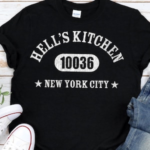 May include: Black t-shirt with the text "HELL'S KITCHEN 10036 NEW YORK CITY" in white. The shirt is paired with blue jeans and white canvas trainers with red and black accents. The items are arranged on a light wood surface.