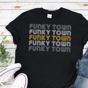 Pode incluir: Camiseta preta com o texto "Funky Town" repetido cinco vezes em estilo retro, com o terceiro "Funky Town" em dourado e o restante em cinza. A camiseta está colocada sobre uma superfície de madeira com um par de tênis brancos e uma calça jeans azul.