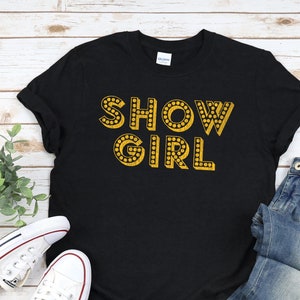 以下が含まれることがあります： 電球のデザインが施された金色の文字で「SHOW GIRL」と書かれた黒いTシャツです。