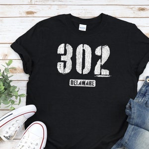 Könnte beinhalten: Schwarzes T-Shirt mit weißem, abgenutztem Text, der "302 DELAWARE" lautet. Das Shirt liegt flach auf einer Holzoberfläche mit einem Paar weißen Sneakers und einer blauen Jeans.