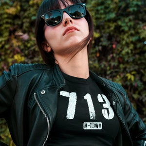 May include: A woman wearing a black leather jacket, a black t-shirt with the number "713" and the text "H-TOWN" printed in white, and a pink and black checkered skirt.